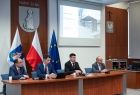 wicemarszałek Łukasz Smółka siedzący na tle flag podczas Forum po obu stronach dwóch innych uczestników obrad