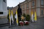 Pomnik Ojca Świętego, przed nim złożone kwiaty i biało-żółte flagi