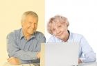 Para seniorów - kobieta i mężczyzna siedzą przy stole przy otwartym laptopie, patrzą przed siebie i uśmiechają się.