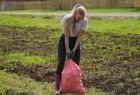 Młoda dziewczyna o blond włosach ubrana w jasną bluzkę, ciemne spodnie i fioletowe gumowce podnosi różowy worek wypełniony ziemniakami