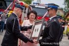 Radna Jadwiga Wójtowicz przekazuje dwóm strażakom z OSP Srebrny Medal Polonia Minor. W tle widoczne wozy strażackie.