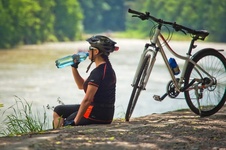 Rowerzystka siedzącą na brzegu rzeki i pijącą wodę z butelki.