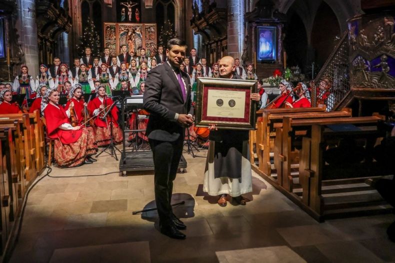 Wicemarszałek Łukasz Smółka przekazuje opatowi medal Polonia Minor. Z tyłu stoi zespół "Mazowsze".