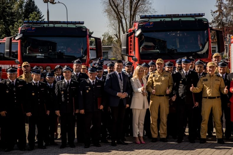 Uczestnicy wydarzenia stoją przed wozami strażackimi.