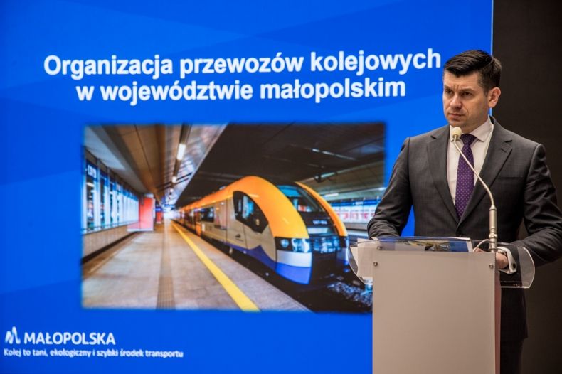 Wicemarszałek Łukasz Smółka przemawia podczas konferencji. Z tyłu widoczna prezentacja i zdjęcie pociągu Kolei Małopolskich.