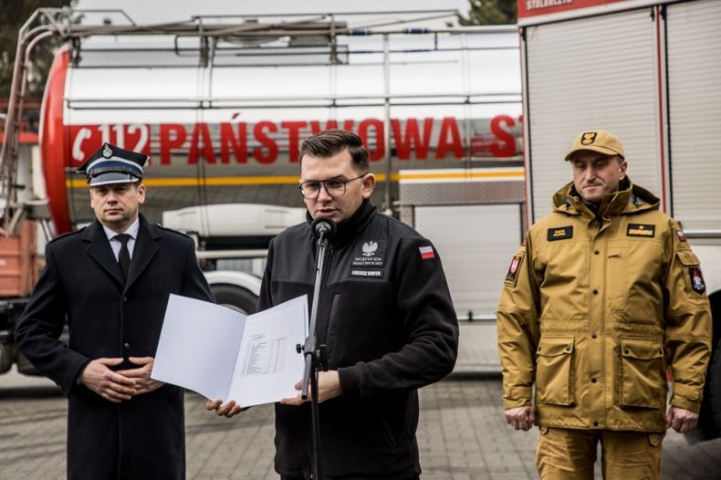 Wicemarszałek Łukasz Smółka podczas konferencji. Z tyłu stoi wóz strażacki.