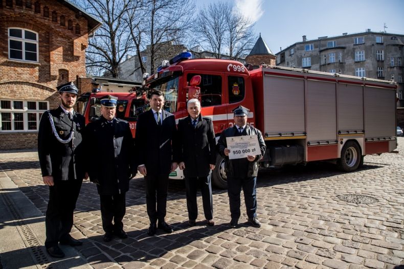 Wicemarszałek Łukasz Smółka stoi ze strażakami. Za nimi widoczny nowoczesny wóz strażacki.