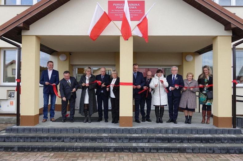 Grupa osób stoi przed budynkiem szkoły i przecina nożyczkami biało-czerwoną wstążkę