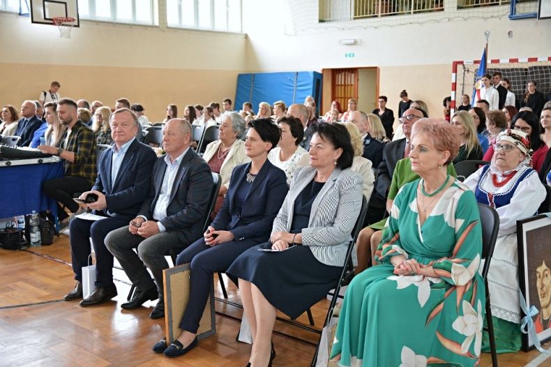 Goście wydarzenia siedzą na krzesłach w rzędach, w tle wnętrze sali gimnastycznej