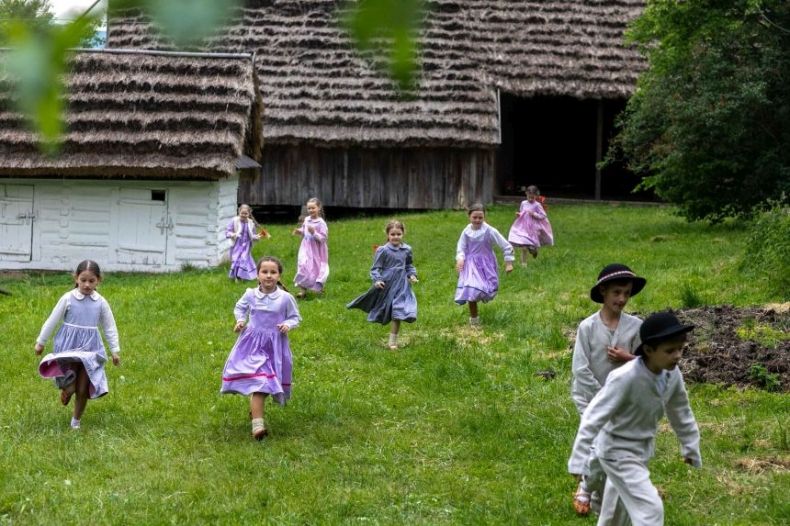Dzieci biegnące po trawie, w tle drewniane budynki kryte strzechą. 