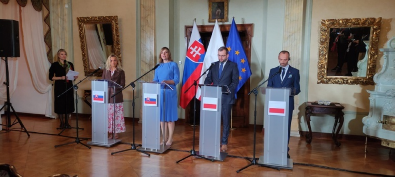 Zdjęcie przedstawia salę z flagami Polski i Słowacji. Po środku przy pulpitach konferencyjnych z godłem Słowacji stoją dwie kobiety, zaś przy pulpitach z godłem Polski dwaj mężczyźni. Osoby te reprezentują rządy obu państw. Obok stoi kobieta - tłumaczka.