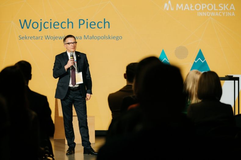 Sekretarz Województwa Małopolskiego Wojciech Piech stoi na scenie i trzyma w ręce mikrofon.