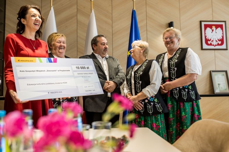 Członek zarządu Marta Malec-Lech trzymająca promesę. Za nią kobiety w strojach regionalnych i mężczyzna w szarym garniturze.