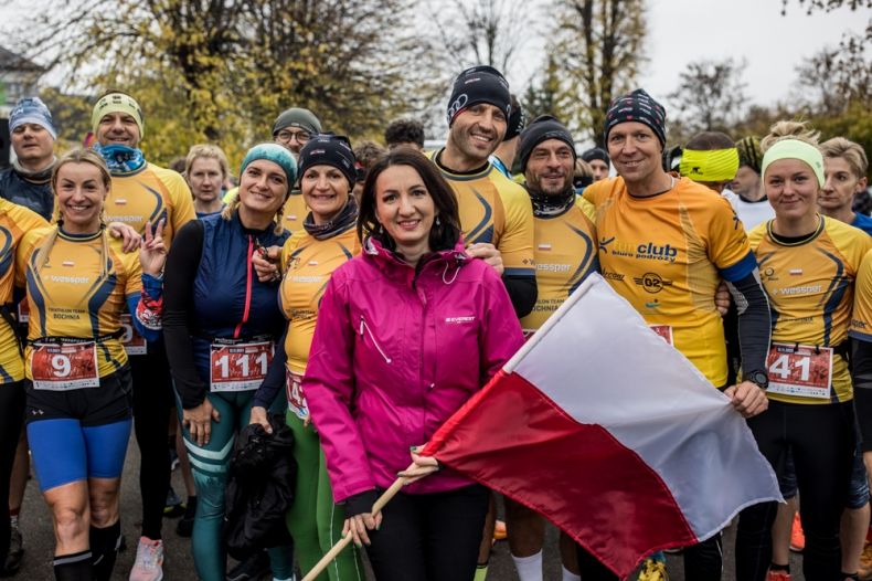 Marta Malec-Lech z zarządu województwa z flagą Polski. W tle widoczni uczestnicy biegu w żółtych strojach.