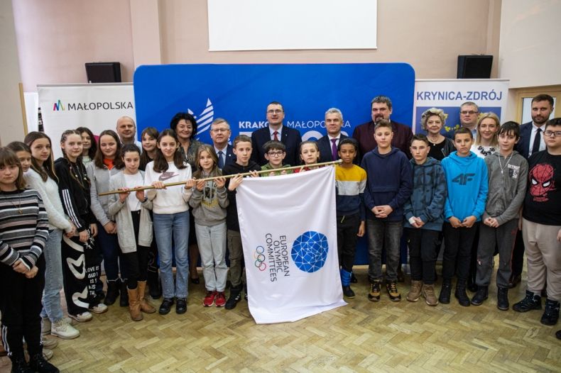 Marszałek Witold Kozłowski, burmistrz Krynicy młodzież, wszyscy pozują wspólnie do zdjęcia z flagą Igrzysk Europejskich. 
