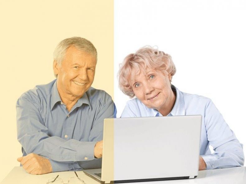 Para seniorów - kobieta i mężczyzna siedzą przy stole przy otwartym laptopie, patrzą przed siebie i uśmiechają się.