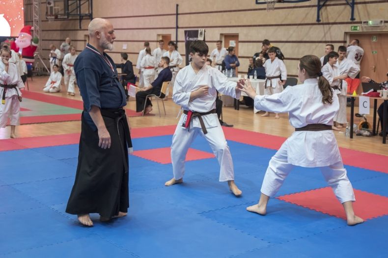 walczą młodzi karatecy, którym sędziuje mężczyzna w tradycyjnym, długim i czarnym stroju 