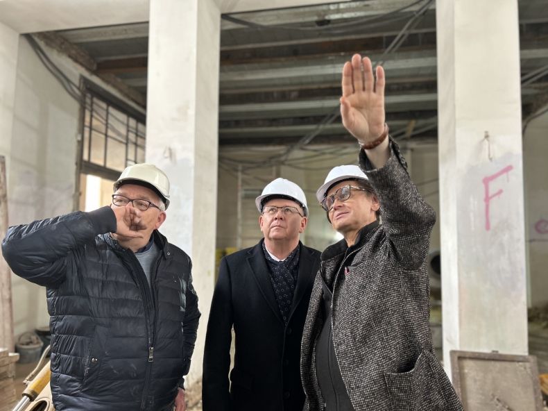 Marszałek małopolski Witold Kozłowski stoi wewnątrz remontowanego budynku. Jeden z mężczyzn stojących po lewej stronie marszałka wskazuję ręką na prace remontowe. 
