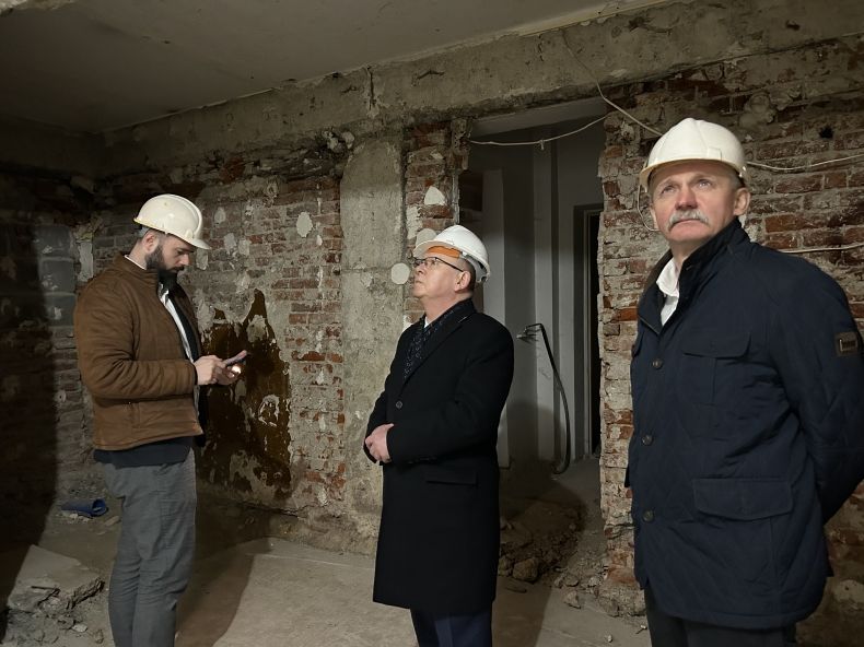 Marszałek Witold Kozłowski wizytuje powstający oddział Muzeum Tatrzańskiego w Zakopanem. Marszałek stoi w towarzystwie dwóch mężczyzn wewnątrz pomieszczenia będącego w surowym stanie. 