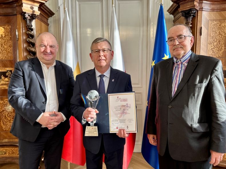 marszałek trzyma dyplom i statuetkę, obok stoją przedstawiciele stowarzyszenia; wszyscy stoją na tle trzech flag: Polski, Małopolski i Unii Europejskiej