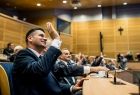 Marszałek Łukasz Smółka siedzi w ławie i podnosi rękę.