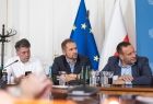 Trzech mężczyzn siedzi przy stole. Za nimi widoczna flaga Polski i Unii Europejskiej.