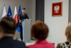 Marszałek Łukasz Smółka stoi przy mównicy. Za nim widoczne flagi, obok godło Polski.