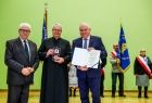 Wicemarszałek Ryszard Pagacz trzyma w rękach dyplom. Obok stoi ksiądz z medalem i mężczyzna.