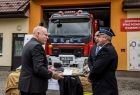 Wicemarszałek Ryszard Pagacz podaje rękę strażakowi. Za nim widoczna remiza i wóz strażacki.