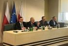 Czterech mężczyzn siedzi przy stole prezydialnym. Za nimi widoczne flagi.