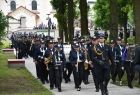 Defilada strażacka idzie wśród zieleni, w tle widoczny kościół.