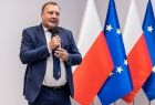 Mężczyzna z mikrofonem stoi na tle flag Polski i Unii Europejskiej.