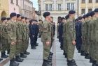 Uczniowie w mundurach stoją w szeregu na rynku.