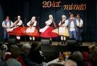 Panie i panowie w strojach ludowych podczas tanecznego występu przed publicznością. Z tyłu, nad sceną, na ciemnej kurtynie znajduje się napis: 20 lat minęło