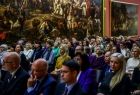 Ludzie siedzą na widowni. Za nim widoczne duże obrazy.