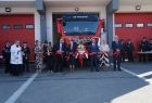 Grupa osób z czerwoną wstęgą w dłoniach przed wozem strażackim.