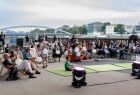 Zdjęcie publiczności na placu Cricoteki. Ludzie - dorośli i dzieci - siedzący na leżakach na placu Cricoteki, w rękach trzymają balony, przed nimi stoją głośniki. W tle widok na Wisłę, Kładkę Bernadkę i drugi brzeg rzeki z budynkami