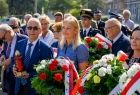 Ludzie stoją na placu i trzymają w rękach wiązanki kwiatów.