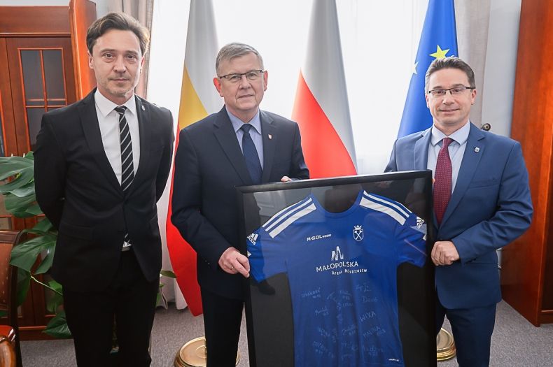 Dwaj mężczyźni oraz wicemarszałek Witold Kozłowski trzymający niebieską koszulkę w ramce.