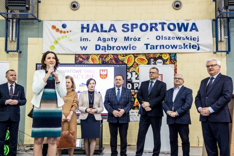 Uczestnicy wydarzenia stoją na hali sportowej.