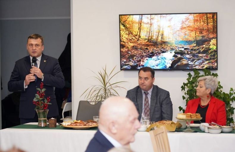 Wojciech Skruch stoi z mikrofonem przy stole. Obok siedzą inni ludzie.