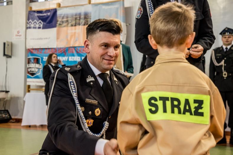 Wicemarszałek Łukasz Smółka ubrany w strażacki mundur rozmawia z chłopcem w stroju strażaka, z napisem straż na plecach.