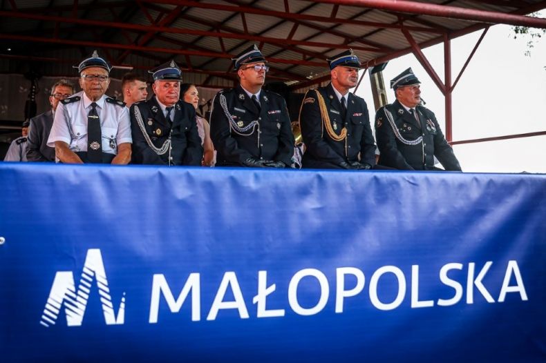 Strażacy siedzą na trybunie honorowej z napisem Małopolska.