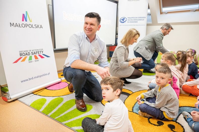 Wicemarszałek Łukasz Smółka przed dziećmi siedzącymi na podłodze. Z tyłu widoczni inni ludzie.