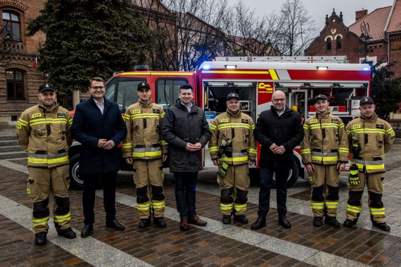 Uczestnicy wydarzenia stoją przed nowym wozem strażackim.
