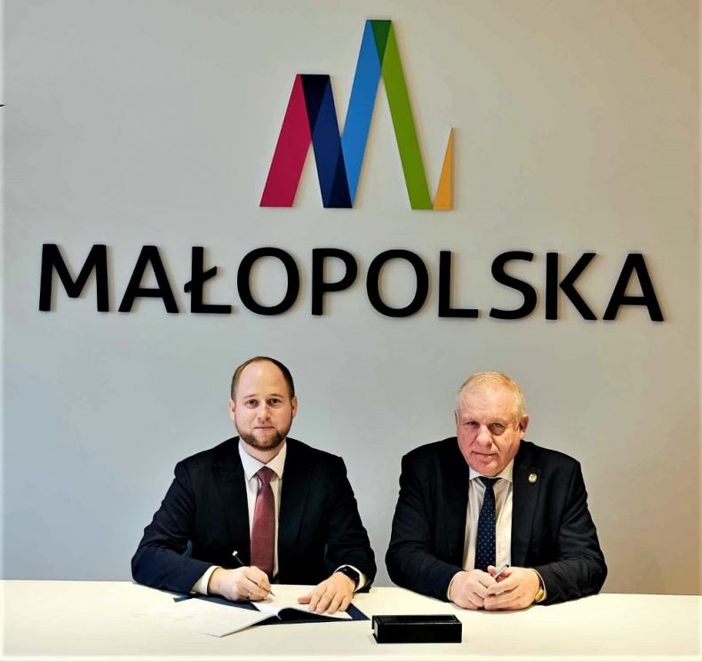 Wicemarszałek Ryszard Pagacz siedzi przy stole. Obok siedzi mężczyzna. Za nimi na ścianie widoczne logo Małopolska.