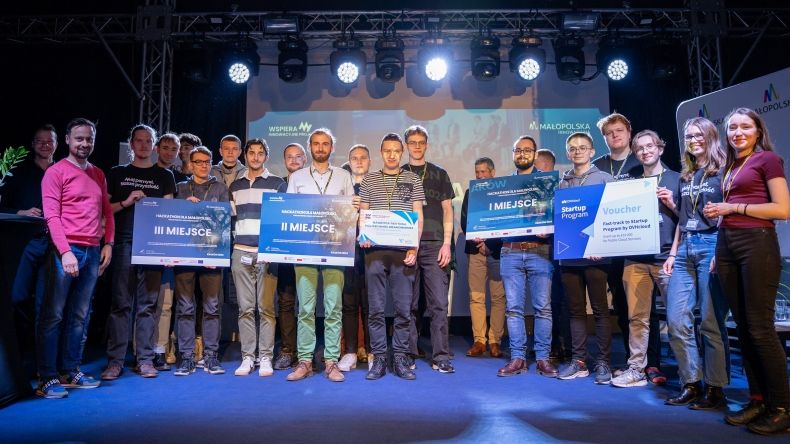 Zwycięzcy Hackathonu stoją na scenie (zespoły). Z góry pada na nich światło. W rękach trzymają tablice z nagrodami