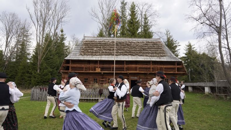 pary w strojach ludowych tańczą przed drewnianą chałupą