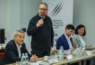 Mężczyzna stoi przy stole. Obok siedzą inni ludzie.