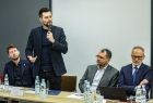 Mężczyzna trzyma w ręce mikrofon. Obok siedzą inni mężczyźni.